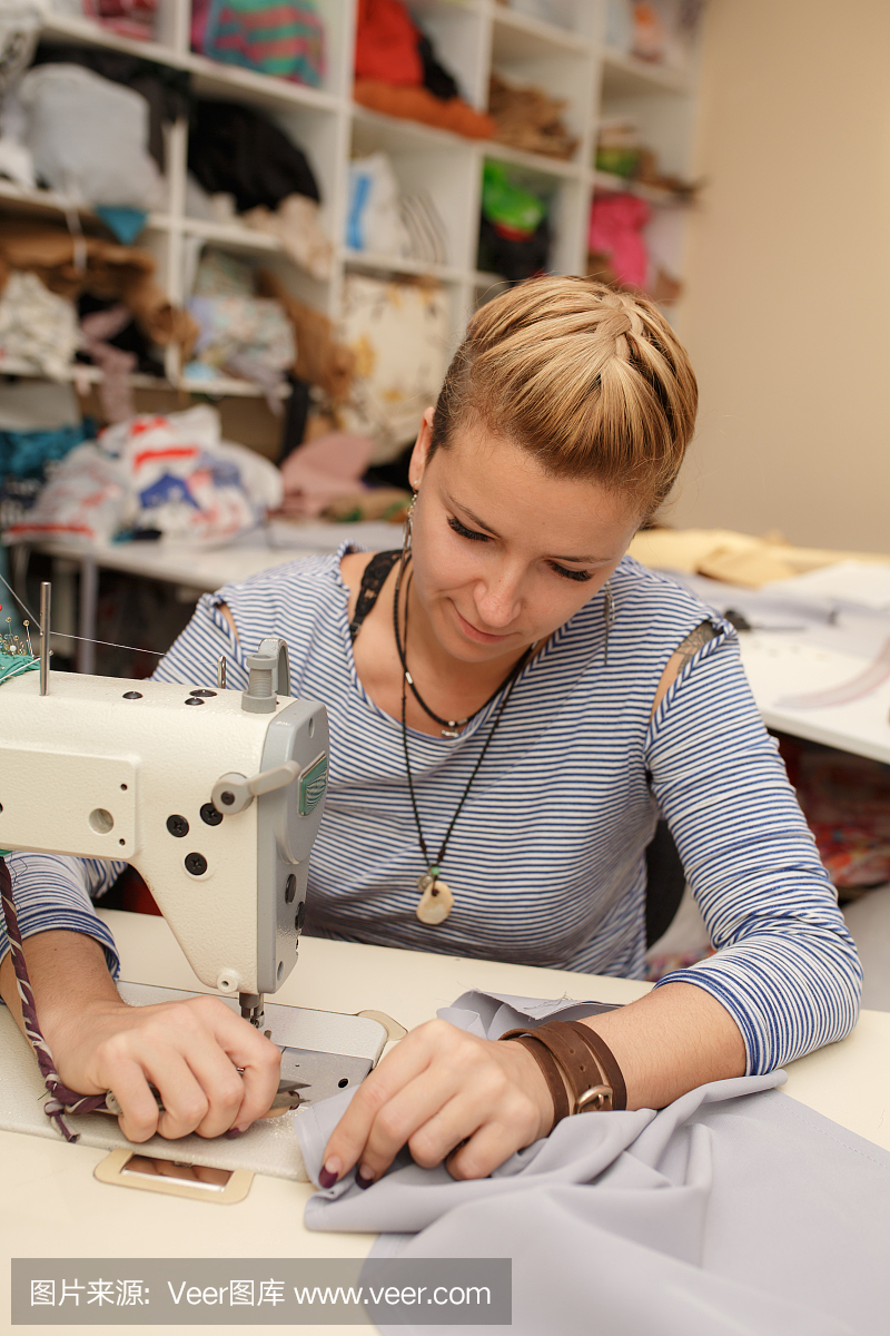 年轻的金发女裁缝布设计师在缝纫机上工作。在她的工作空间。服装制造业。